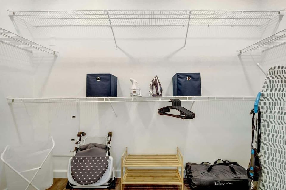 A walk-in-closet with plenty of shelves, hangers, iron & board, shoe rack, crib, high chair, and vacuum
