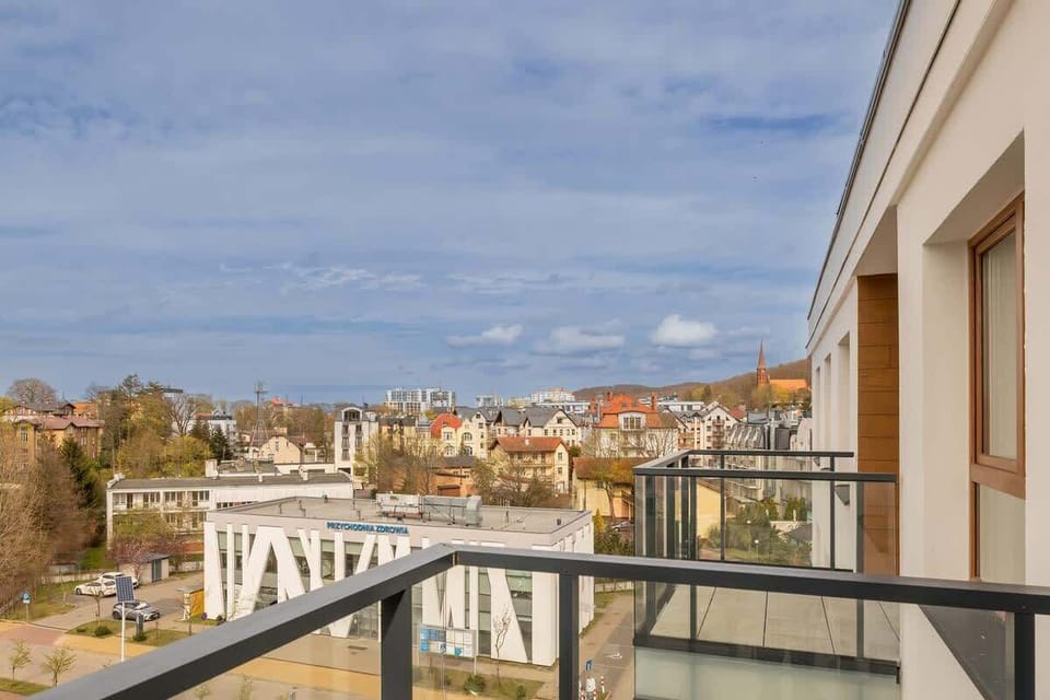 A panorama from the balcony, capturing rooftops, trees, and distant sea under a partly cloudy sky.


