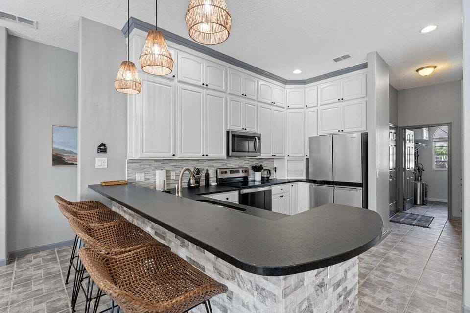 Modern kitchen with stainless steel appliances, sleek black countertops, and a large island with bar seating. Stylish wicker lighting and bar stools add a warm, coastal touch. Perfect for cooking and gathering.