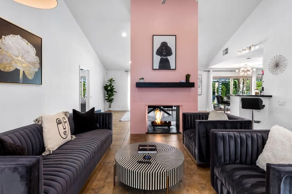 Main living room - lit fireplace on pink accent wall, velvet sofas, striped oval coffee table, vaulted ceilings. Resort energy, every night.