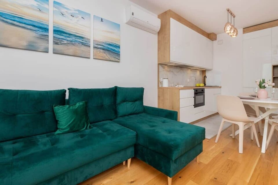 A modern living room with a green sofa, coffee table, and a dining area in the background. The space is open and bright.