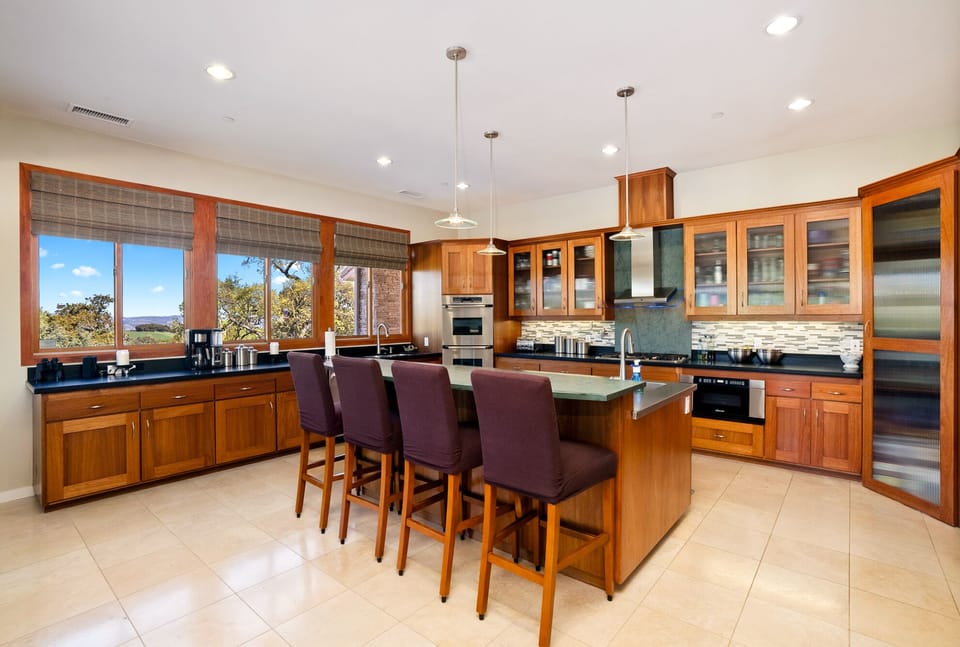 This professional chef's kitchen features rich wood cabinetry, a mosaic backsplash, and a large island with elegant purple barstools. Modern stainless steel appliances and pendant lights complete this high-end space.