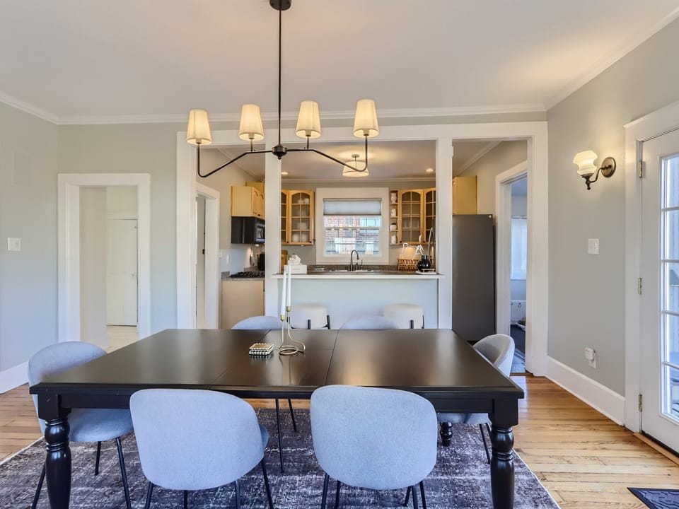 A modern dining room with a black table, six light gray chairs, and a chandelier. The dining area opens to a kitchen with light wood cabinets, granite countertops, and stainless steel appliances.