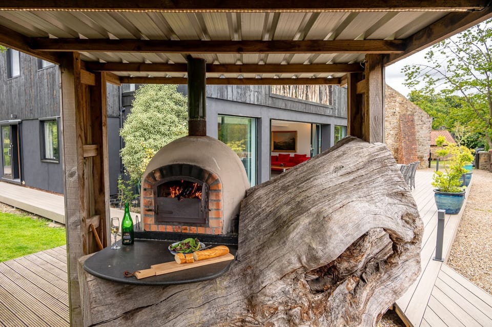 Outdoor wood-fired pizza oven and covered cooking area beside a modern countryside cabin terrace.