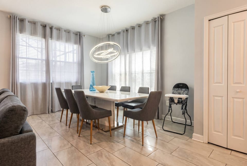 This inviting dining area boasts a sleek white table and stylish gray chairs, ideal for family meals or gatherings, bathed in soft natural light from large windows that highlight the warm tones of the contemporary tile flooring.