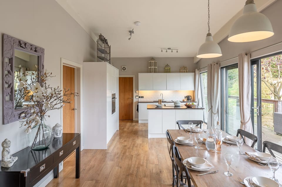 Open-plan kitchen and dining area at Abbey, with a central island, dining table for six, and large doors leading to the lakeside deck.