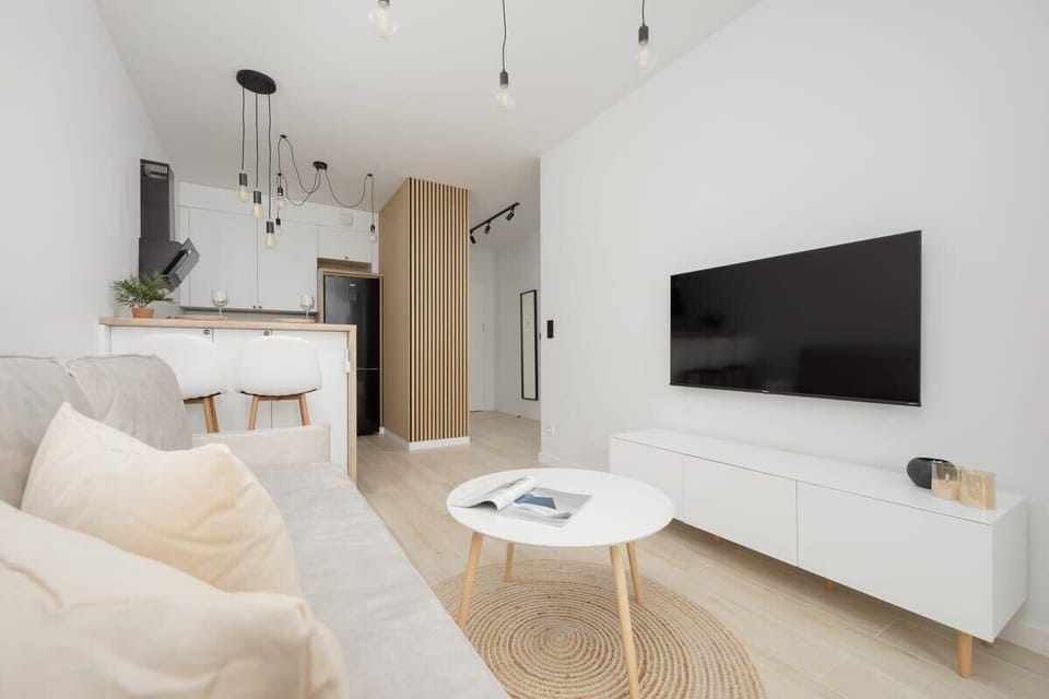 A spacious and bright living room with neutral tones, a soft rug, and a dining table in the background. A versatile space for both lounging and dining.