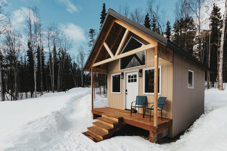 Tiny cabins exterior featuring a charming wooden wall, inviting white door, and two cozy chairs for enjoying the surrounding beauty.