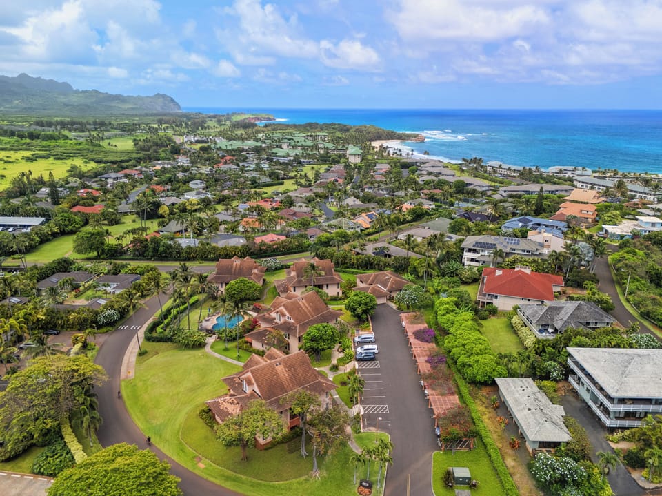 Aerial view of a coastal residential community nestled between lush mountains and the sparkling Pacific Ocean, showcasing the property's stunning tropical Hawaiian setting.