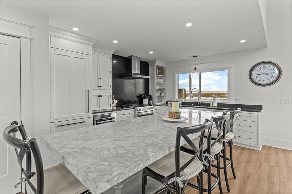 Bright kitchen with a large island, barstools, black countertops, white cabinets, open shelves, stainless range hood, farmhouse sink, and wood-look floors.