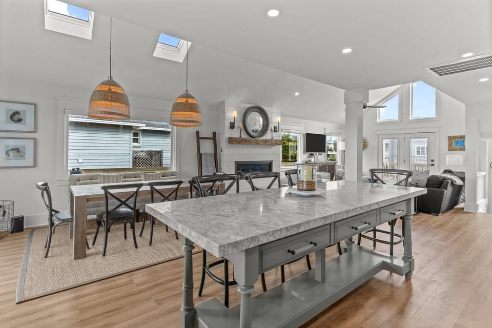 Open dining and living space with a large table, woven pendant lights, fireplace, vaulted ceilings, tall windows, French doors, and bright coastal seating.