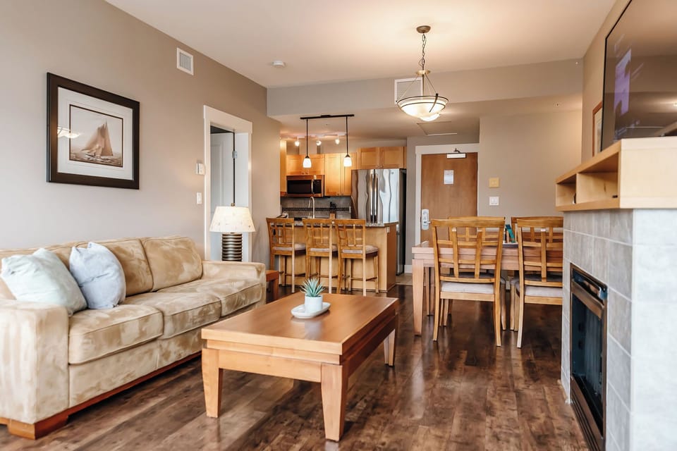 This open-concept condo features a tan couch with pillows, a wooden coffee table, and a dining table for six. The kitchen has wood cabinets, light countertops, and a breakfast bar. A stone fireplace with a TV adds a cozy touch to the space.