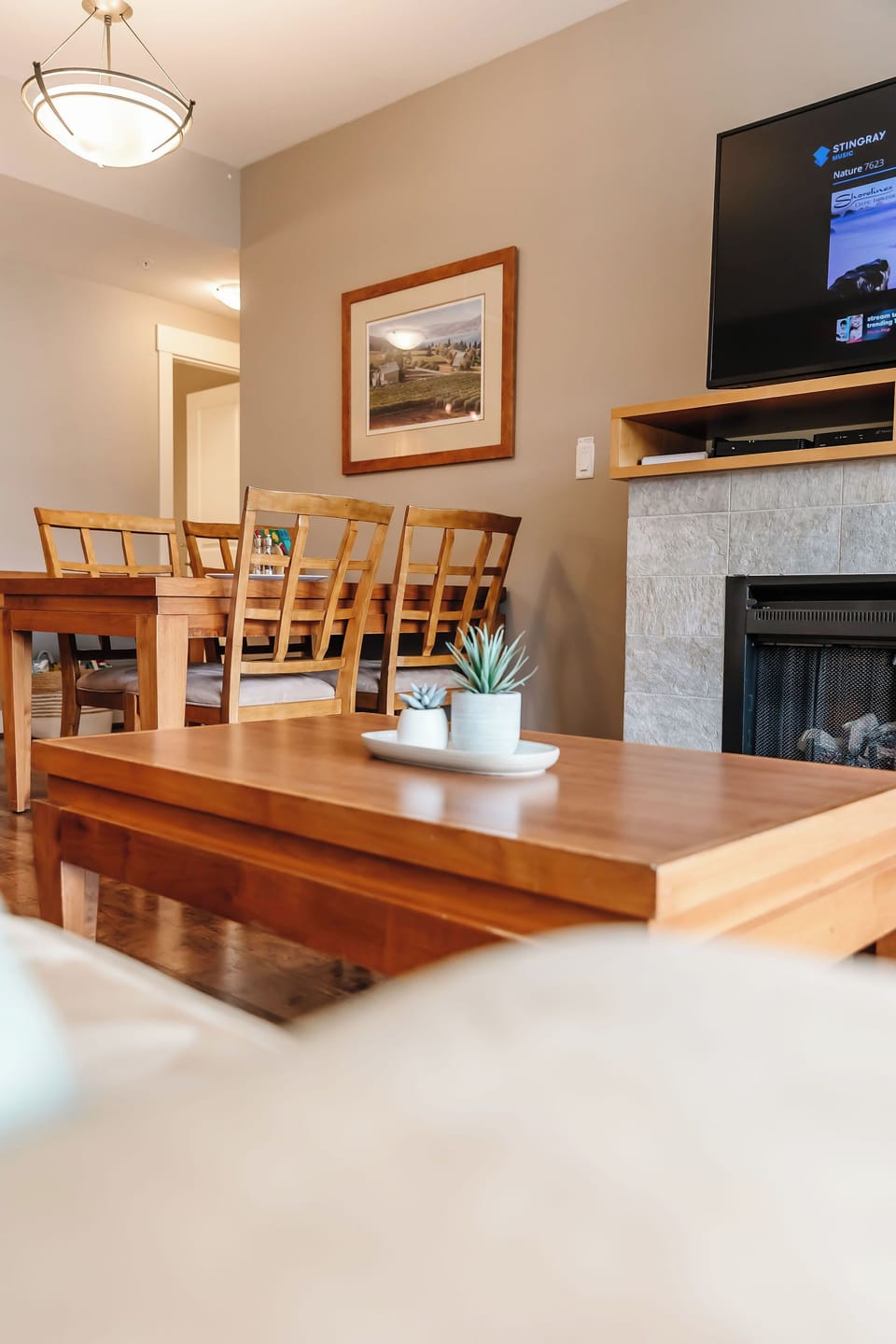 This cozy living and dining room features a white couch, wooden coffee table with plants, and a stone fireplace with a mounted TV. The dining area has a wooden table with six chairs. Hardwood floors and neutral walls complete the inviting space.