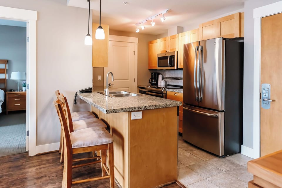This stylish kitchen features stainless steel appliances, granite countertops with a light brown and black pattern, and light wood cabinets. The island with beige cushioned bar stools adds a modern touch.