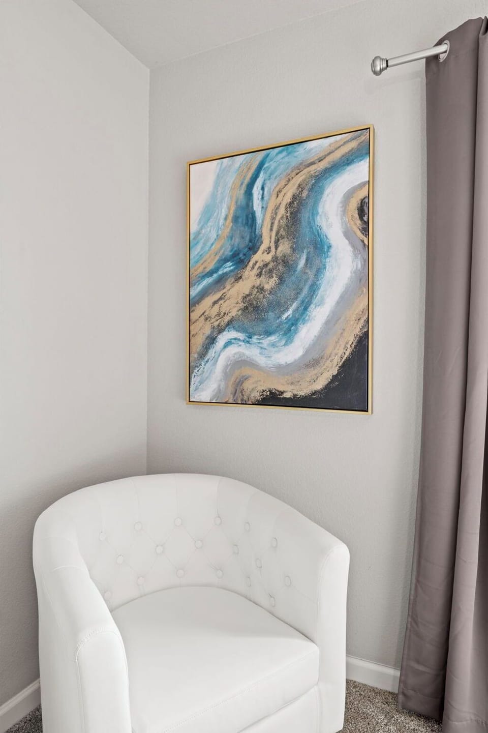 A tufted white armchair sits in the corner of a room with gray walls, next to a window with gray curtains. A framed abstract painting with blue, white, and gold swirls hangs on the wall.