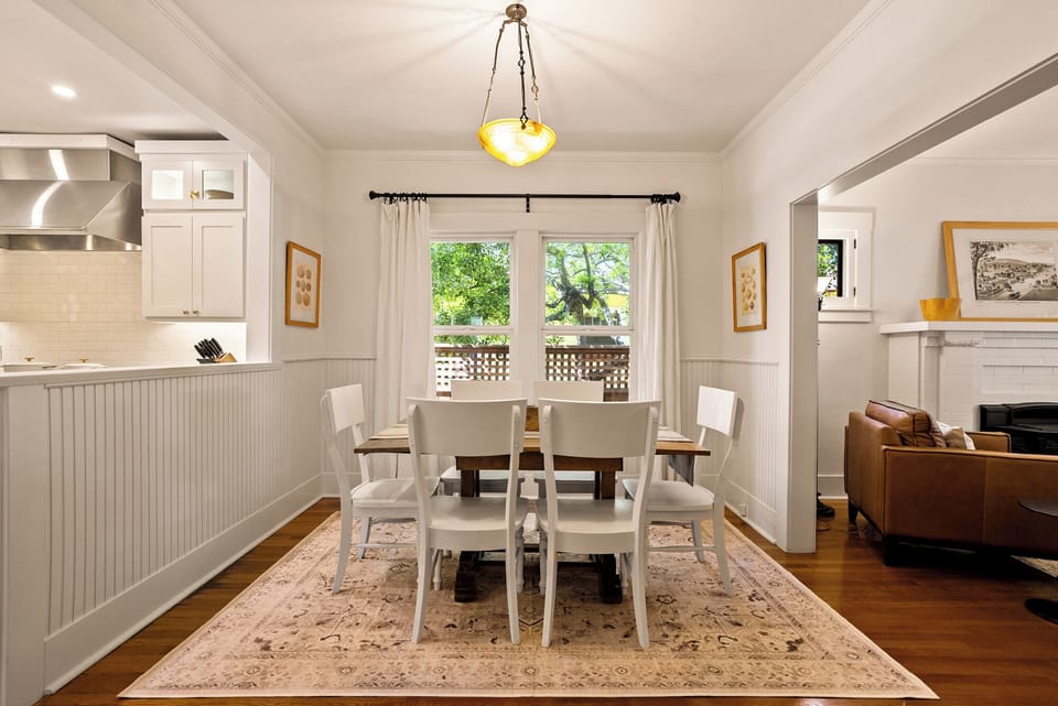 An elegant formal dining area, offering a classic wood table paired with crisp white chairs, set atop a sophisticated patterned area rug, and beautifully defined by white wainscoting and large windows that provide a view of the lush greenery.