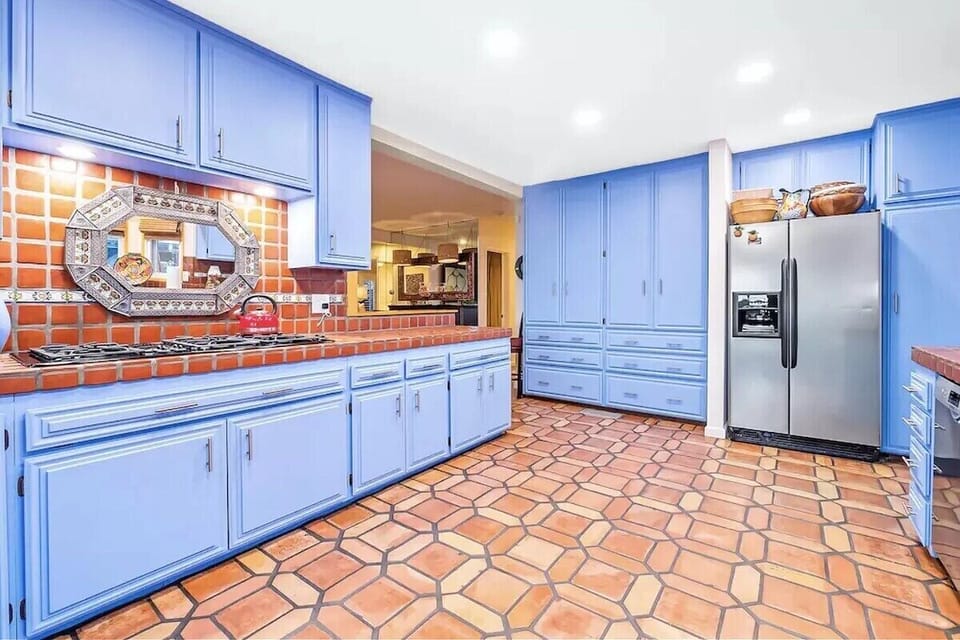 Spacious kitchen with vibrant blue cabinets, warm terracotta tile flooring, stainless steel refrigerator, intricate tile backsplash, decorative mirror, ample storage, and a cozy Southwestern-inspired aesthetic.