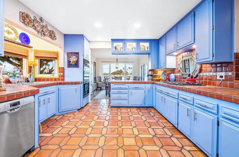 Bright and spacious Mediterranean-style kitchen with striking blue cabinetry, terracotta tile floors, tiled countertops, stainless steel appliances, decorative accents, and an open layout leading to a waterfront dining area.