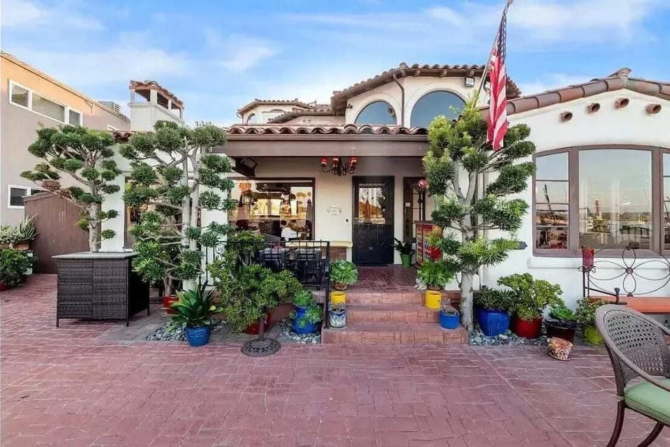 Stunning Spanish-style home with terracotta roof, arched windows, lush greenery, vibrant potted plants, cozy outdoor seating, elegant black door, decorative lighting, and a charming brick-paved courtyard.