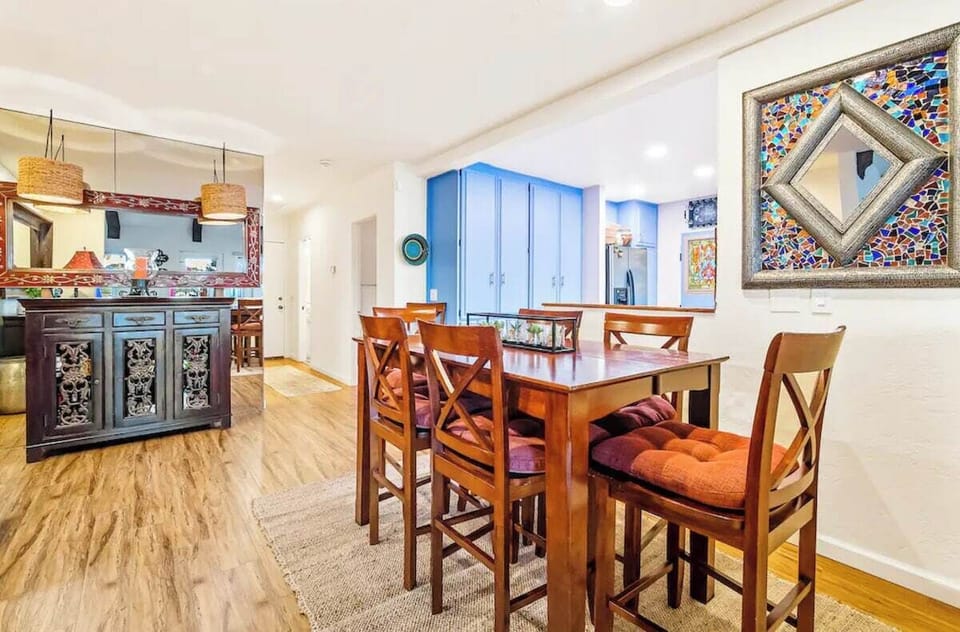 Elegant dining area with warm wood furniture, plush seat cushions, stylish mosaic mirror, sleek hardwood floors, and an open layout leading to a vibrant kitchen with blue cabinetry and a charming bar with pendant lighting.