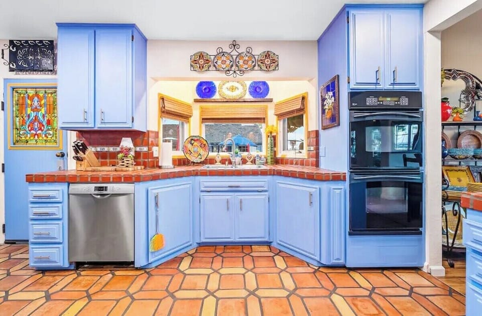 Vibrant kitchen with sky-blue cabinets, terracotta tile flooring, colorful decor, tiled countertops, stainless steel appliances, double ovens, and a charming backsplash, creating a warm and inviting culinary space.