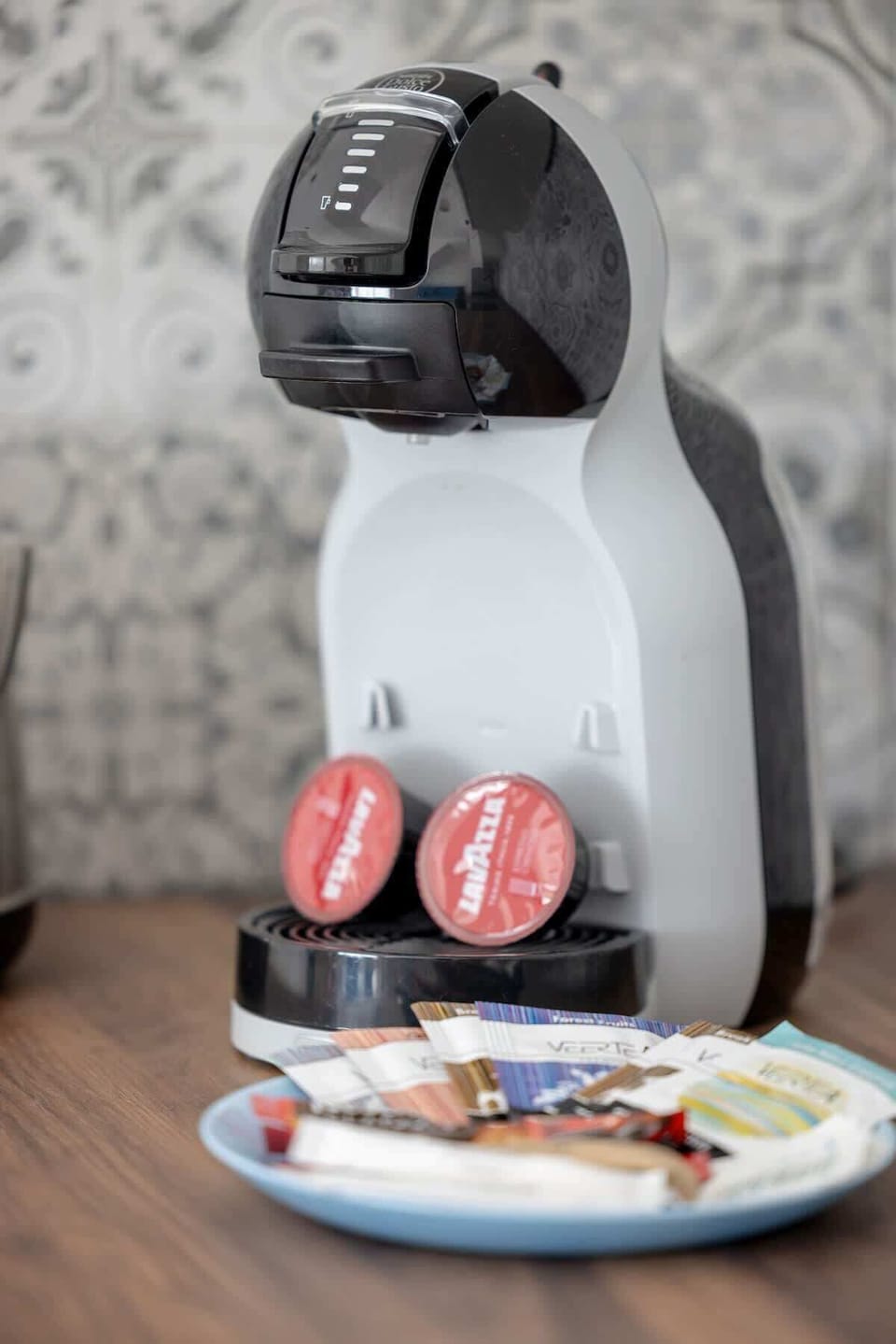 A capsule coffee machine with two red pods and a small plate against a patterned wall creates a cozy coffee corner in the apartment.