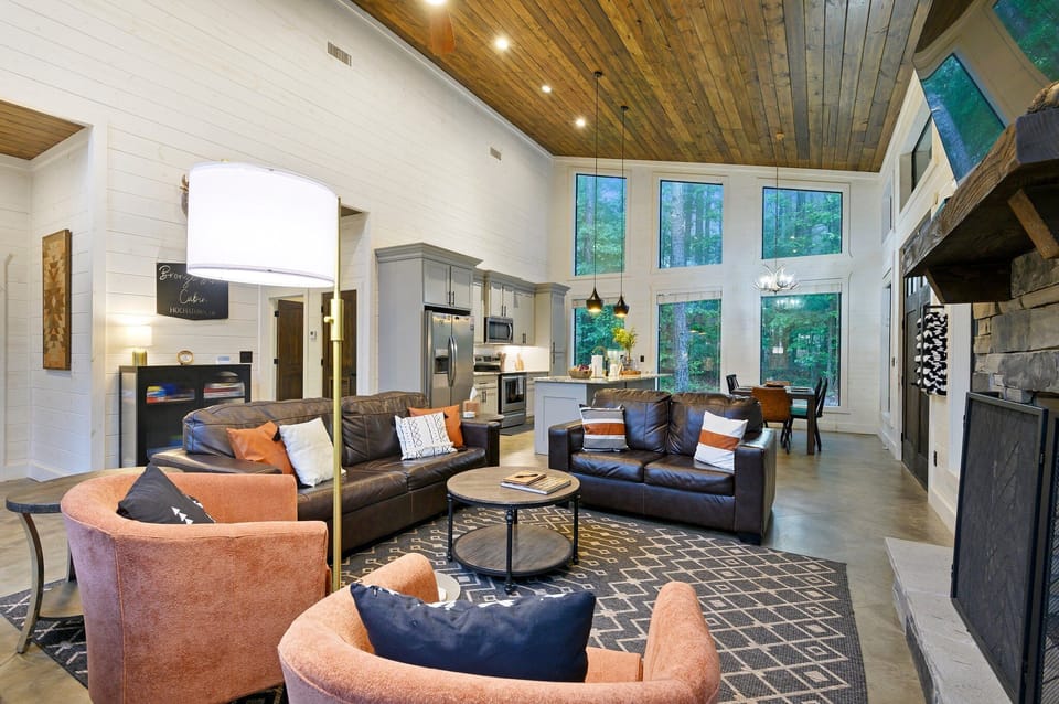 Spacious living room with large windows, wooden ceiling, and modern furnishings including brown sofas, orange chairs, a round coffee table, and a fireplace. The open kitchen and dining area are visible.