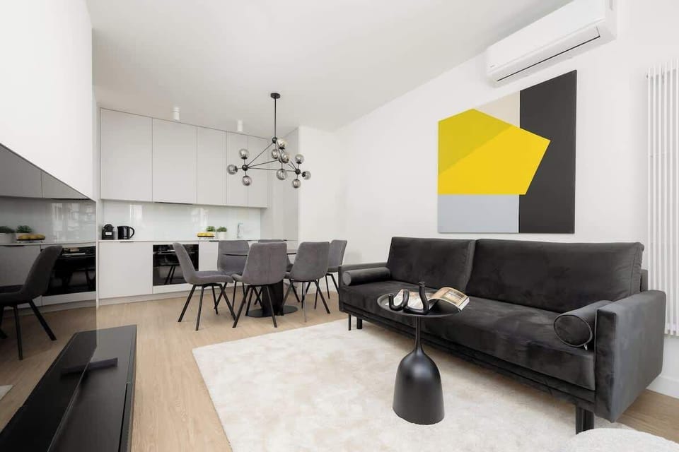 A bright living room combining open kitchen and lounge space, furnished in monochrome tones with splashes of yellow.