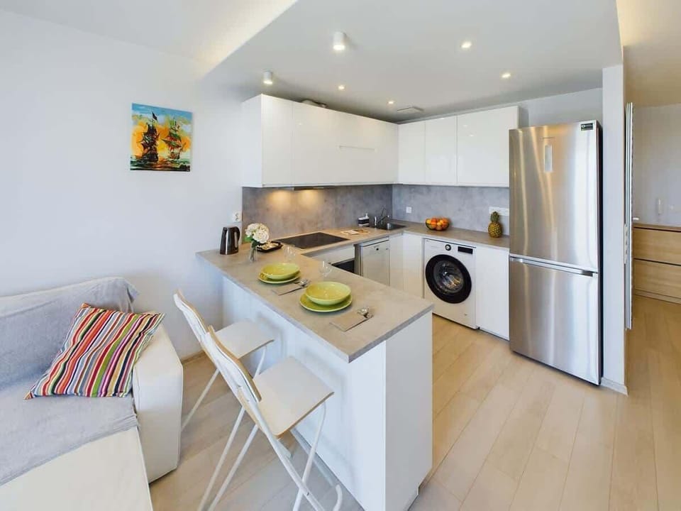 A kitchen and dining combo with a small table for two, fridge, and modern cabinets in bright lighting.