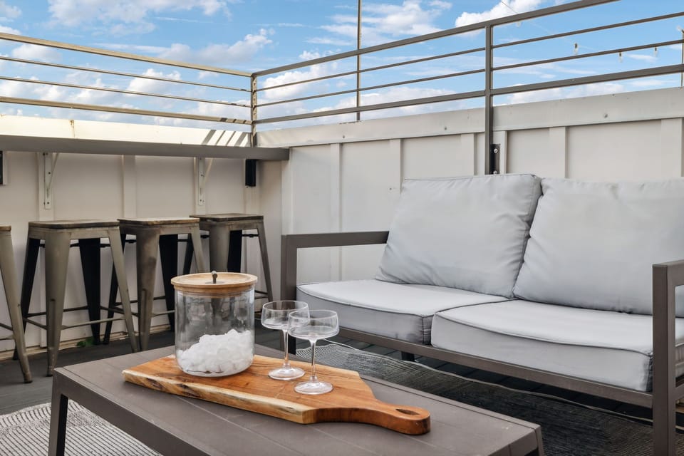 An outdoor patio with a sofa, a table holding a jar with a lid, two glass goblets, and a wooden tray. In the background are several high stools along a railing under a cloudy sky.