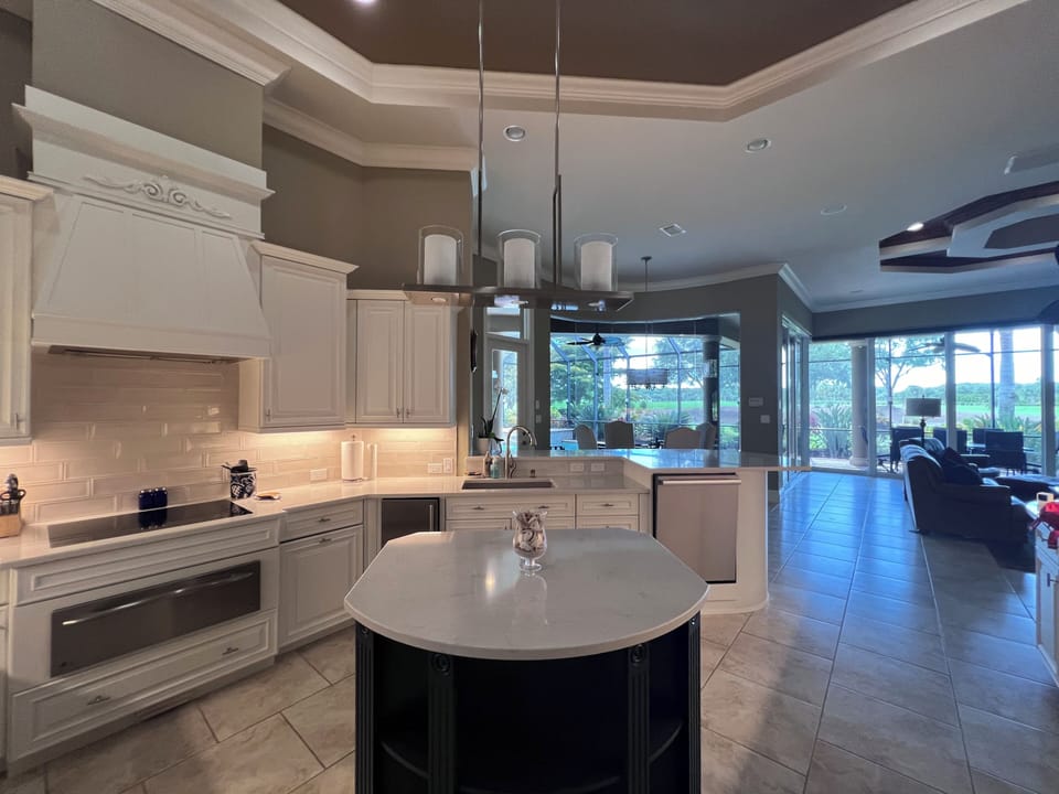 A spacious modern kitchen with a central island, white cabinets, stainless steel appliances, and pendant lighting. Beyond the kitchen, a living area with large windows overlooks a green outdoor view.