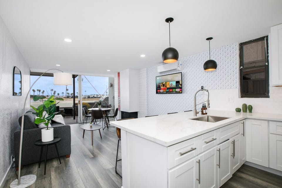 A modern and airy open-plan living area featuring a pristine kitchen with a large island, contemporary pendant lights, and sleek white cabinetry. The space flows seamlessly into a cozy living and dining area, with expansive glass doors offering breathtaki