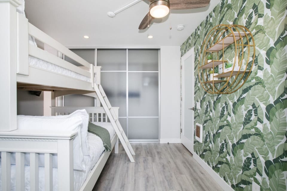 A bright and cheerful bedroom featuring a bunk bed with crisp white linens, a modern ceiling fan, and stylish sliding closet doors. The room is adorned with vibrant tropical wallpaper and a unique circular shelf, creating a fun and inviting space perfect 