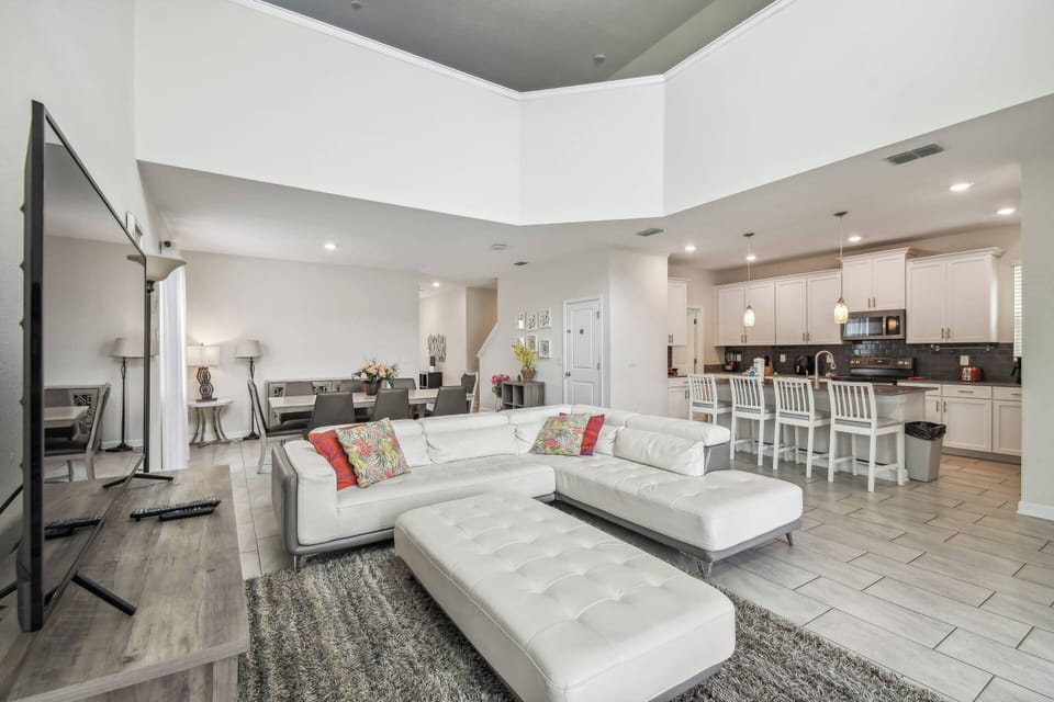 Experience a bright and airy living area with high ceilings and a contemporary white sectional, perfect for relaxation. The stylish decor, warm accents, and cozy rug enhance the open layout, flowing beautifully into the kitchen.