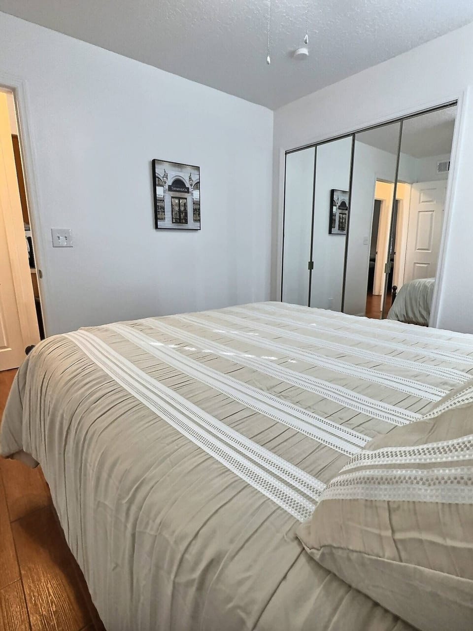 Welcome to this cozy bedroom featuring a spacious plush bed with a stylish striped blanket. Enjoy warm wooden flooring, a mirrored closet for added openness, and soft lighting that creates an inviting atmosphere for your stay.