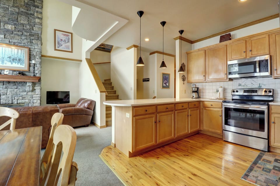 A modern kitchen and living area featuring wooden cabinets, stainless steel appliances, a stone fireplace, and leather furniture. A staircase leads to the upper floor.