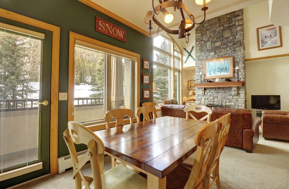 A cozy dining area with a wooden table and chairs is adjacent to a living room with a stone fireplace. Large windows offer a snowy outdoor view of the mountain.