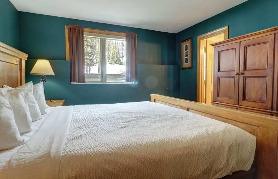 A cozy bedroom features a wooden bed frame with white linens, a wooden dresser, and a bedside lamp. The room has teal walls, a window with brown curtains, and a door leading to another space.