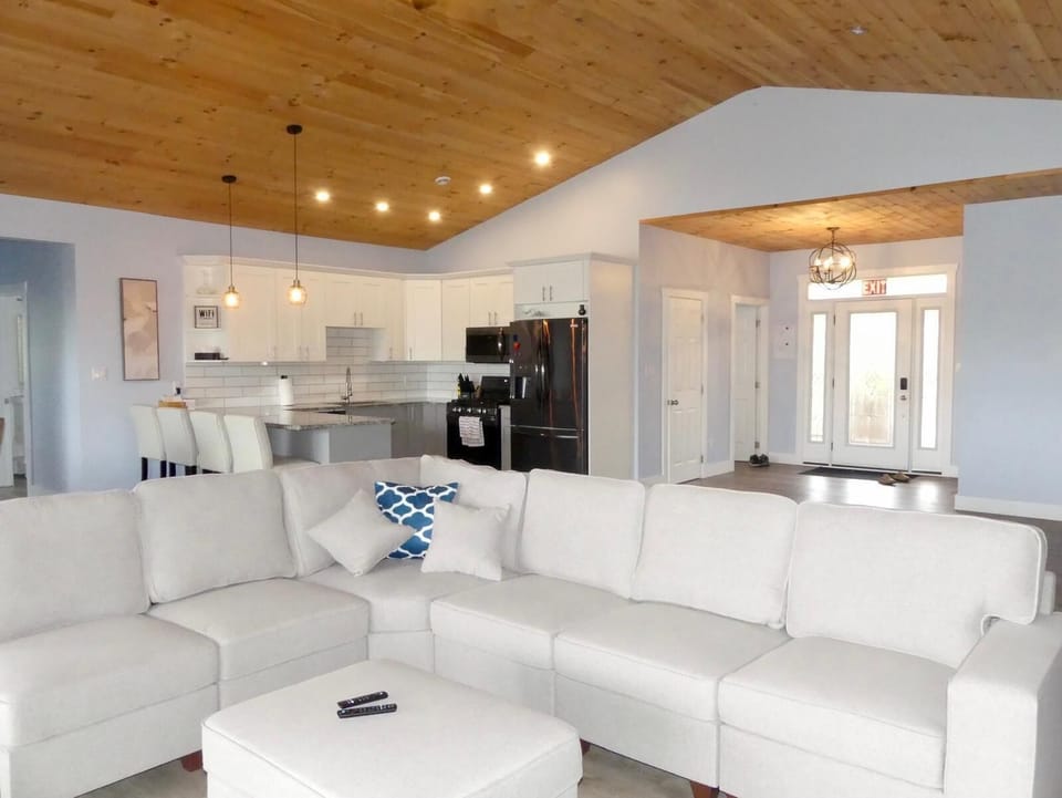 Inside, the expansive open-plan living area is bathed in natural light. The high, wood-paneled ceiling and large windows create an airy, inviting space, perfect for family gatherings and entertaining friends.