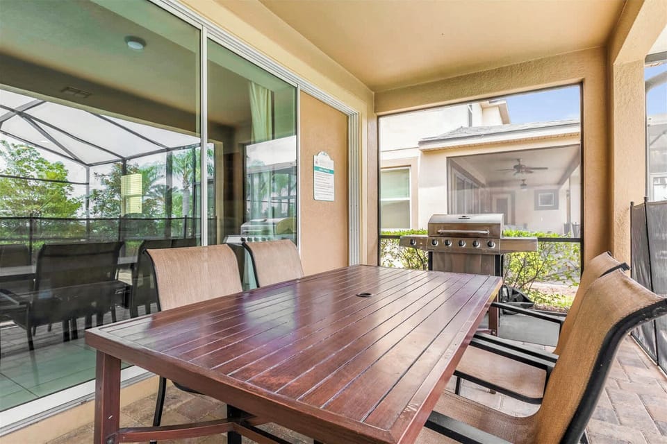 Covered patio dining area with a spacious wood table, cushioned seating, and stainless-steel grill for relaxed outdoor meals.