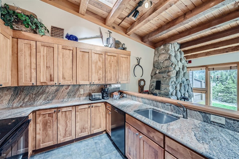 A rustic kitchen with wooden cabinets, stone countertops, built-in appliances, and a large stone fireplace. Exposed wooden beams adorn the ceiling. A window offers a view of greenery outside.