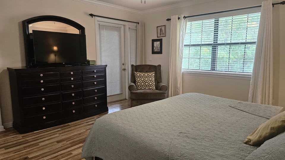Bedroom with wood flooring, a dark dresser with mirror, cushioned armchair, and window with blinds and curtains.