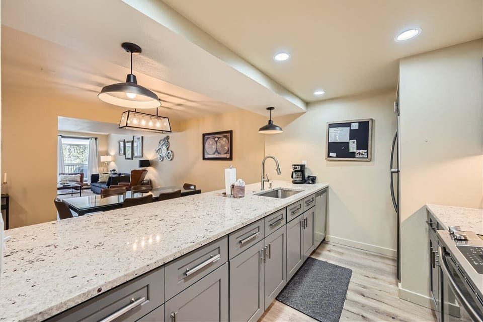 A modern kitchen with gray cabinets, white granite countertops, pendant lighting, a sink, and stainless steel appliances. The kitchen opens to a dining area and living room with hardwood flooring.