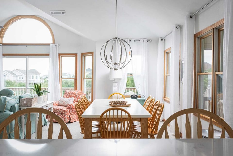 Bright Dining Area w/ Long White Table, Natural Wood Chairs, Modern Globe Chandelier, Large Windows & Open-Concept Kitchen Access