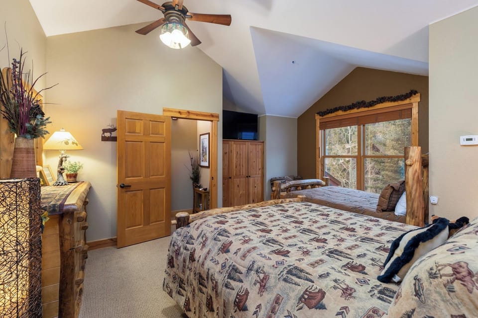 A rustic-style bedroom features two beds with patterned covers, wooden furniture, a ceiling fan, a window with a nature view, and warm lighting.