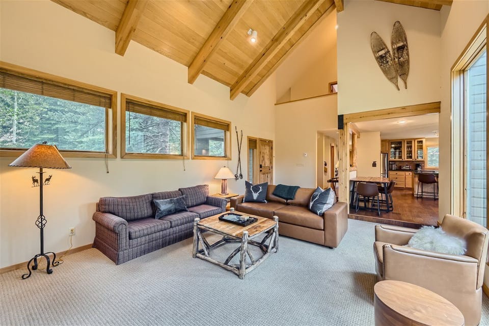 A rustic living room with vaulted wooden ceiling, large windows, two sofas, a small armchair, a coffee table, and a standing lamp. The open layout extends to a dining area and kitchen in the background.