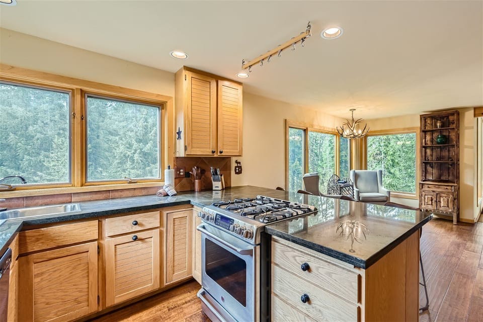 A modern kitchen with wooden cabinets, a central island with a stove, and large windows overlooking a forest. Adjacent dining area with a chandelier and glass table.
