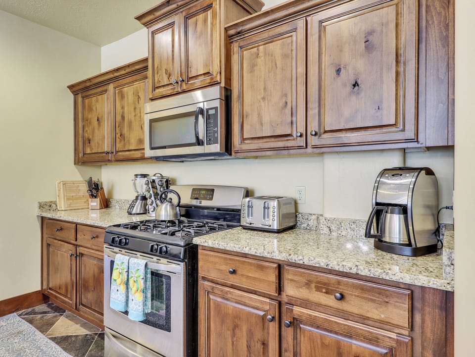 View of fully equipped kitchen featuring a coffee maker, microwave, stove/oven, upgraded cabinetry, granite countertops, blender, kettle, cooking spices, and stainless steel refrigerator