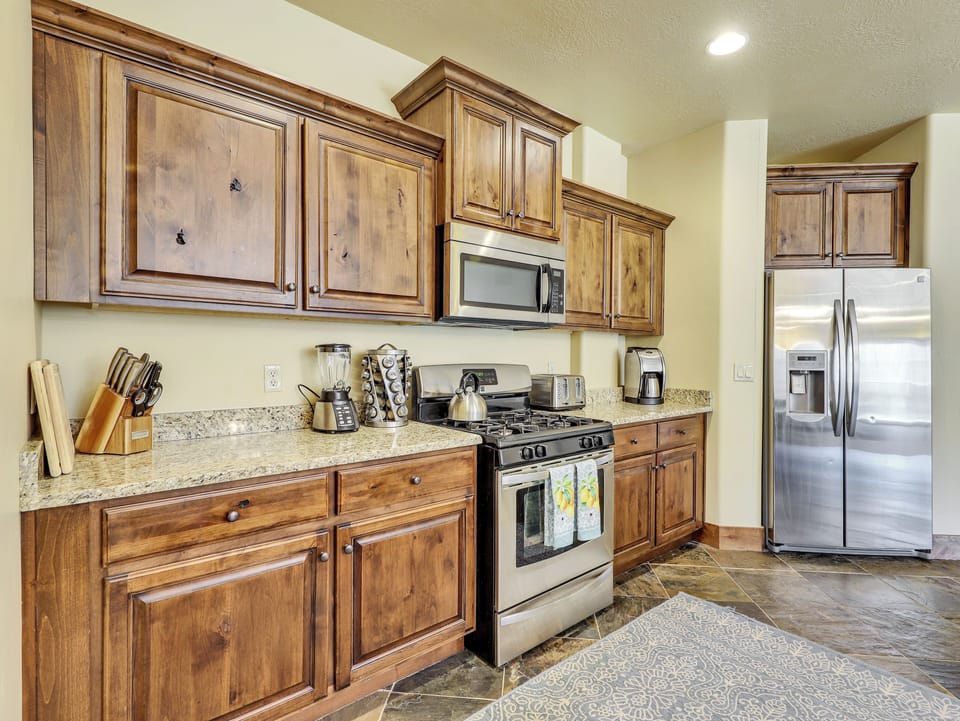 View of fully equipped kitchen featuring a coffee maker, microwave, stove/oven, upgraded cabinetry, granite countertops, blender, kettle, cooking spices, and stainless steel refrigerator