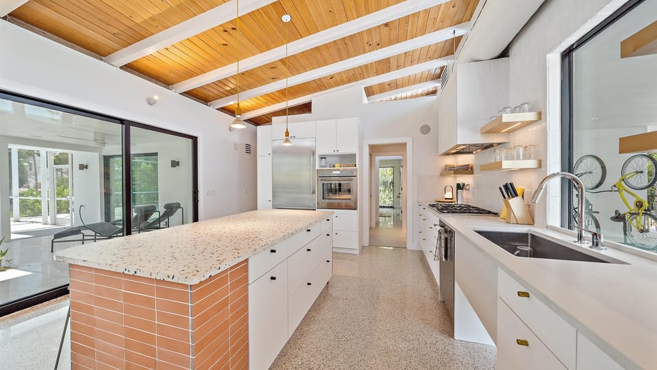 Modern kitchen with white cabinets, a large island with a terrazzo countertop, stainless steel appliances, and wooden ceiling beams. A sliding glass door leads to an enclosed porch area.